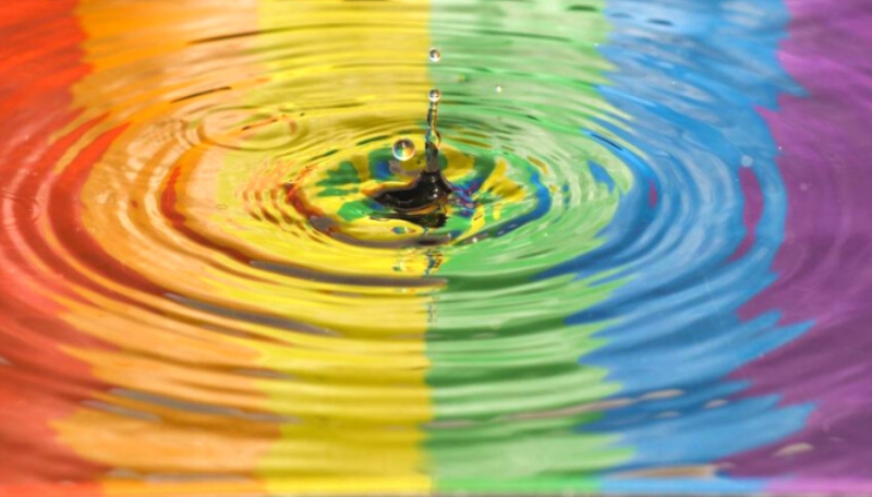 Ein Wassertropfen tropft in eine Wasserpfütze, in der sich eine Regenbogenflagge spiegelt.