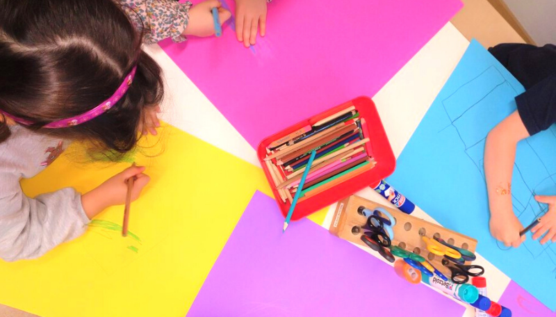 Auf einem Tisch liegen Buntstifte und Papier in den Farben pink, gelb und blau. Zwei Kinder malen auf dem Papier.