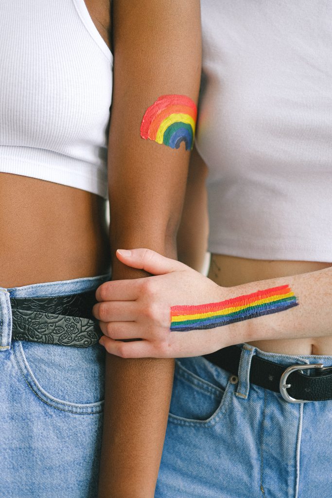 Zwei Jugendliche stehen nebeneinander. Sie tragen beide blaue Jeans und weiße, bauchfreie T-Shirts. Auf ihren Armen sind bunte Regenbögen, als Zeichen der LGBTQ+ - Szene, mit Schminke gezeichnet. 