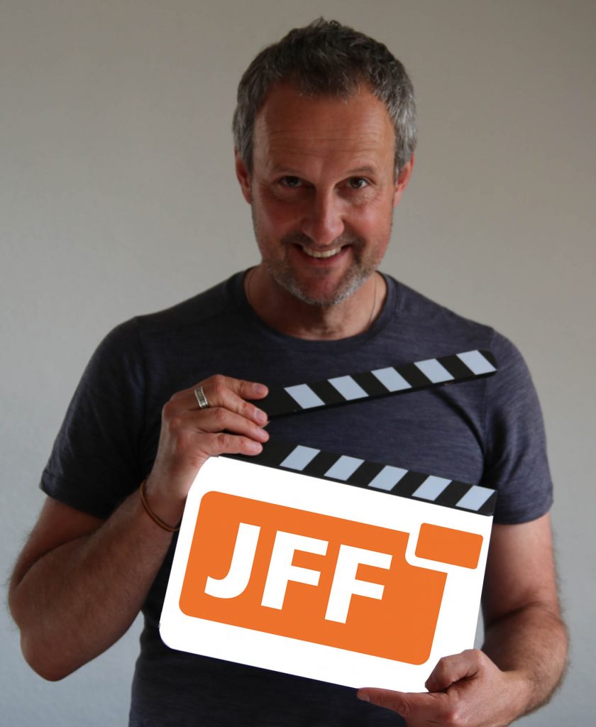Frank Fineiß hat eine Filmklappe in den Händen, auf der das JFF-Logo ist. Er grinst in die Kamera.