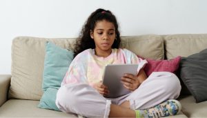 Kind sitzt auf einem Sofa und hält ein Tablet in der Hand. Sie schaut fragend auf den Bildschirm.