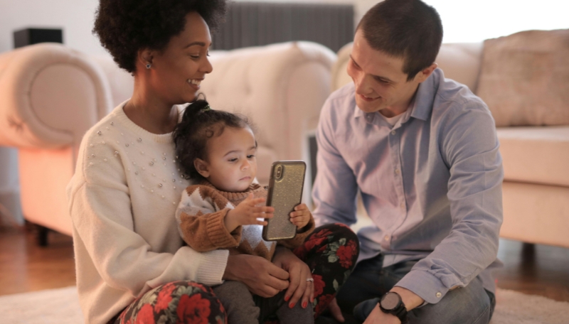 Eltern mit Kind sitzen gemeinsam auf dem Teppich und schauen in ein Smartphone.