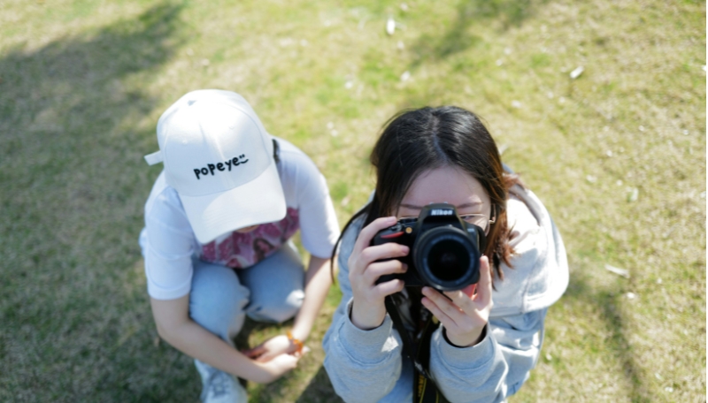 2 Jugendliche. Person eins Sitzt auf der Wiese, man sieht nur die Cap. Person 2 steht daneben und fotografiert nach oben. man sieht das Gesicht nicht, aber die Kamera