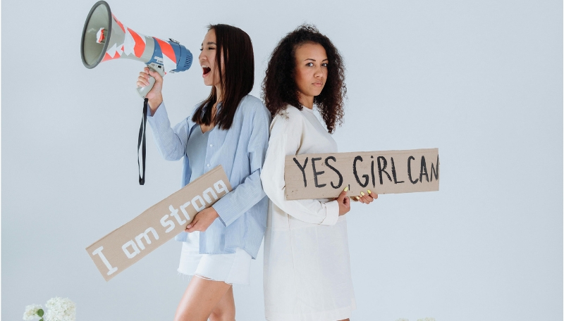 zwei weiblichgelesene Personen, die Rücken an Rücken stehen. Die linke hat ein Megaphone und ein Schild "I am strong" in der Hand, die rechte Person ein Schild mit "Yes, Girl can"