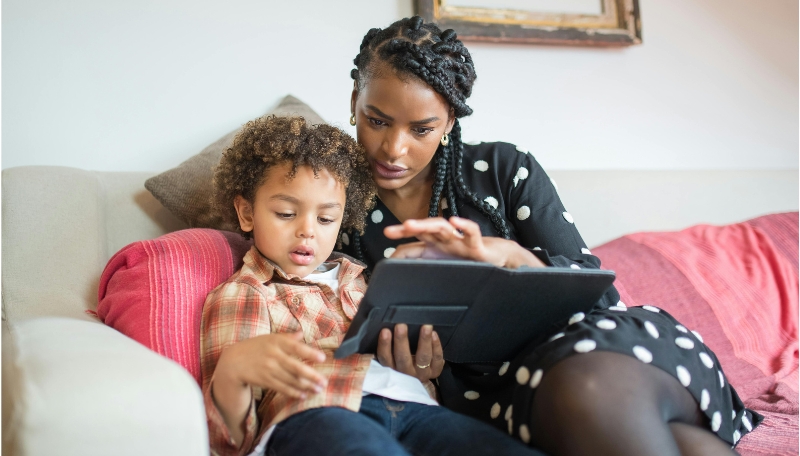 Kind und eine erwachsene Person sitzen auf einem Sofa und machen etwas gemeinsam am Tablet