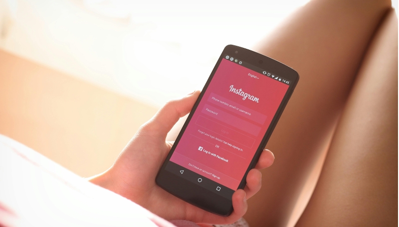 Person hält Smartphone in der Hand. Man sieht die linke Hand und Oberschenkel der Person. Auf dem Smartphone ist die Anmeldeseite von Instagram zu sehen